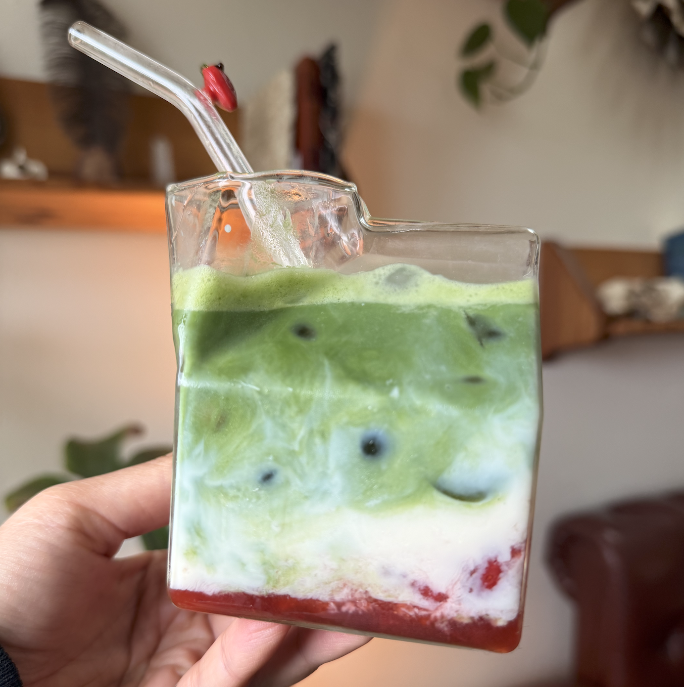 Hand holding a square glass of iced strawberry matcha latte with vibrant layers of red syrup, oat milk, and bright green matcha